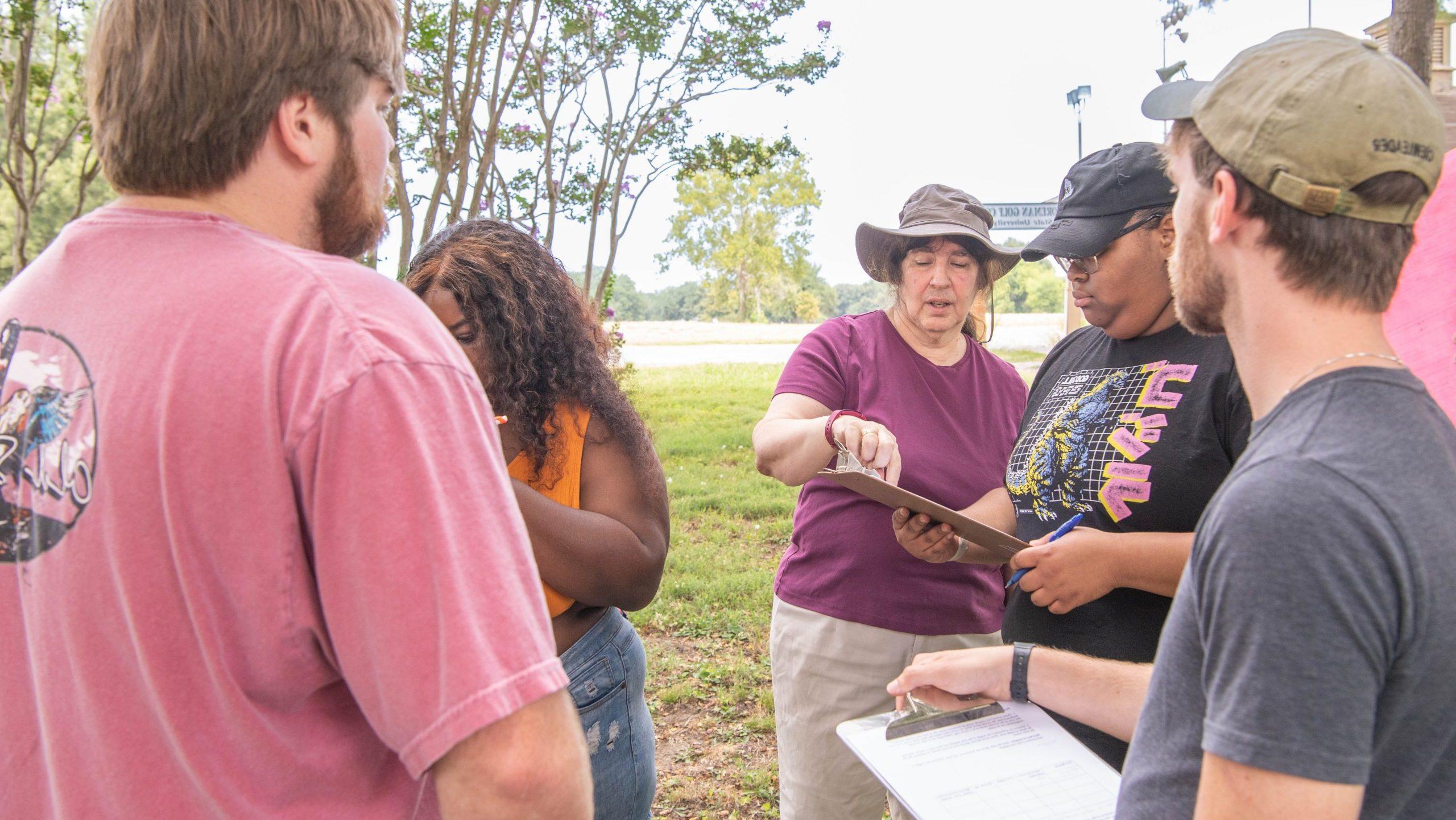 Professor with four students outside writing and reviewing data on clipboards.